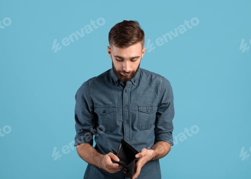 Preview: Poverty and unemployment. Desperate young man showing empty wallet with no money over blue studio