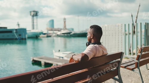 Preview: Young african guy relaxes sitting on bench in the port on yachts and ships background. Back view