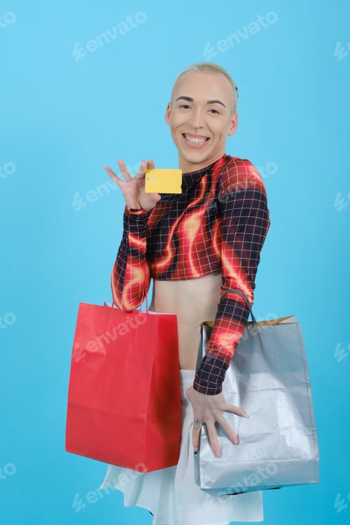 Preview: Young smiling non-binary person showing a credit card to the camera while holding shopping bags