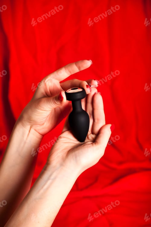 Preview: Holding Black Sex Toy in Front of a Red Fabric Background in the Studio