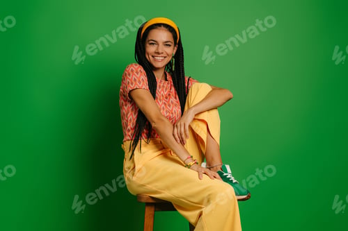 Preview: Beautiful woman with dreadlocs looking at camera while sitting on the chair against green background