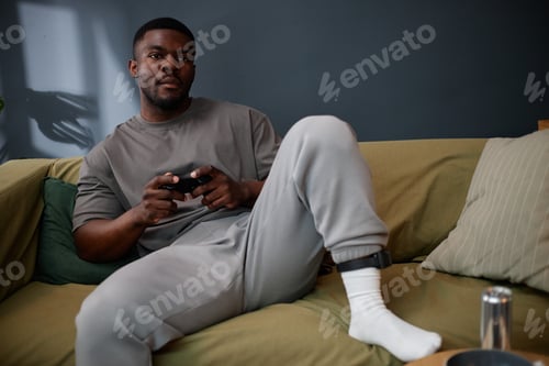 Preview: Young Adult Black Man Sitting on Sofa Wearing Ankle Monitor Playing Video Game
