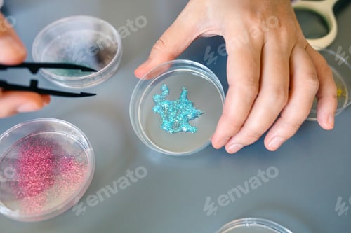 Preview: Close up of female scientist hand holding a blue glitter sample over petri dish on laboratory