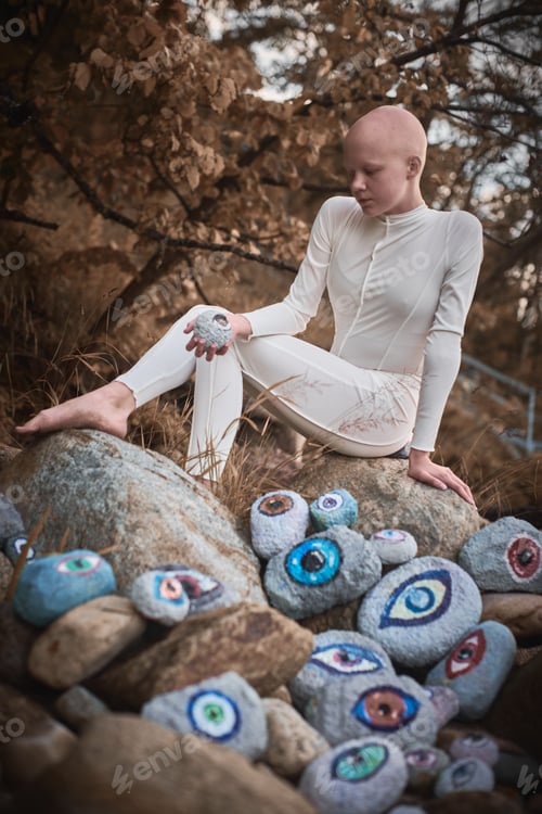 Preview: Young hairless girl with alopecia in white futuristic costume pensively examines stone with eye