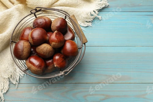Preview: Roasted edible sweet chestnuts in metal basket on light blue wooden table, top view. Space for text