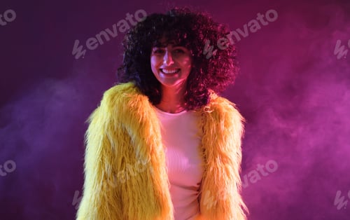 Preview: Beautiful young woman in yellow fur coat on color background in neon lights and smoke