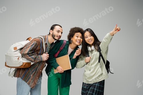 Preview: cheerful asian woman looking away and pointing with finger near multiethnic students isolated on