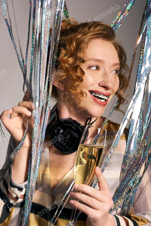 Preview: Joyful young woman gift wrapping with sparkling decorations and holding a glass in a festive setting