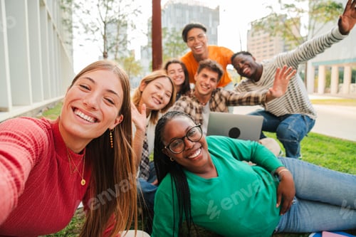 Preview: Blonde female student smiling taking a selfie with her classmates sitting at university campus lawn