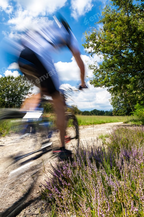 Preview: Cyclist in blurred motion riding on a rural road through green spring meadow