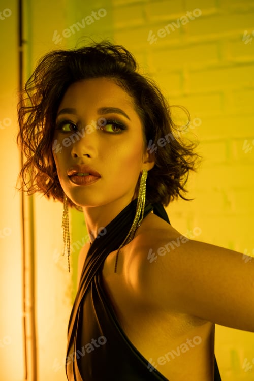 Preview: Young asian woman in earrings and dress looking away while standing in neon light in night club