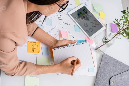 Preview: Young female designer making notes in copybook while bending over documents