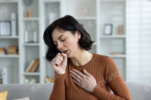 Preview: Close-up photo of a young woman at home, sitting on the couch and coughing hard, feeling chest pain