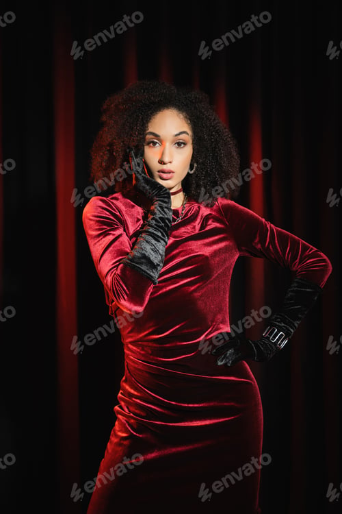 Preview: Stylish african american woman in burgundy dress and gloves standing near red drapery