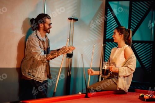 Preview: Young couple talking while playing billiard and drinking beer in pool hall.