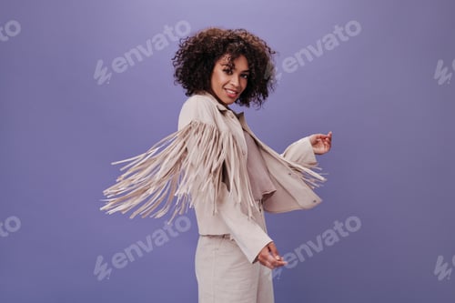 Preview: Woman with wavy hair dances on purple background. Brunette curly woman in beige outfit posing and m