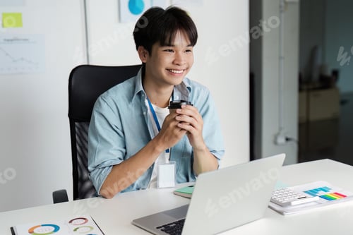 Preview: Smiling young man enjoying coffee while working on a laptop in a modern office.