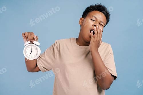 Preview: Portrait of tired African American teenager holding alarm, yawning, wake up in the morning