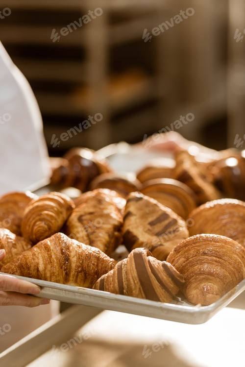 Preview: cropped shot of female baker holding tray with croissants on baking manufacture