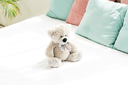 Preview: grey teddy bear near pink and blue pillows on white bedding in bedroom