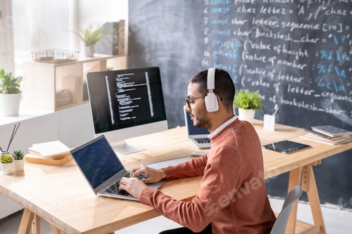 Preview: Busy young programmer with headphones analyzing data on laptop display
