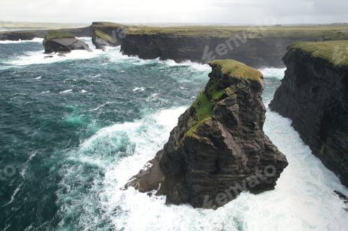 Preview: Dramatic cliffs of Kilkee, Ireland, with waves crashing against rugged rocks under a cloudy sky