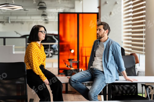 Preview: Bearded Hispanic man in casual denim sitting relaxed on the office table, talking with his colleague