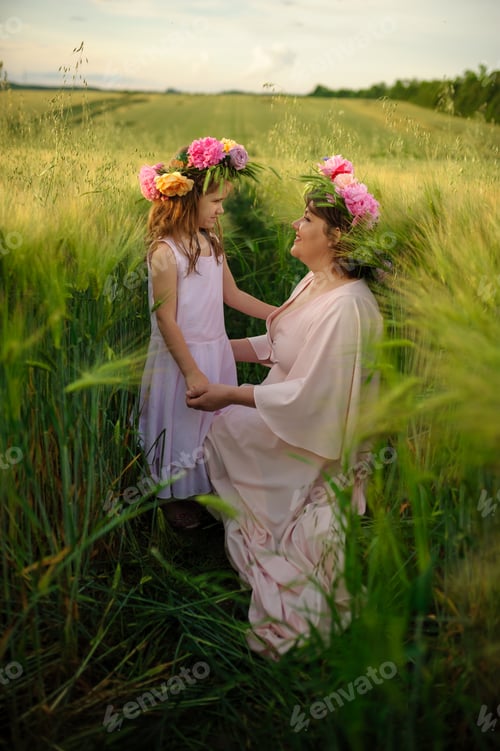 Preview: Mother and daughter in pink dresses in a green wheat field. On their heads they have floral wreaths.