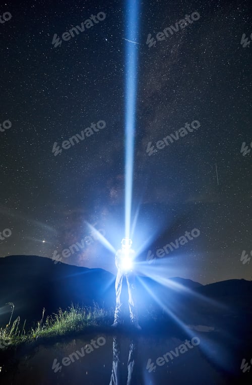 Preview: Spaceman directs a ray of light into the sky with a help of a lantern in the night in the mountains