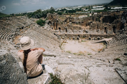 Preview: A teenage girl in a hat sits on top of an amphitheater in Perge, Turkey. Back view