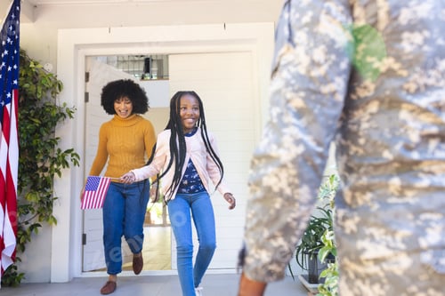 Preview: Happy african american wife and daughter welcoming male soldier outdoors with usa flag, copy space