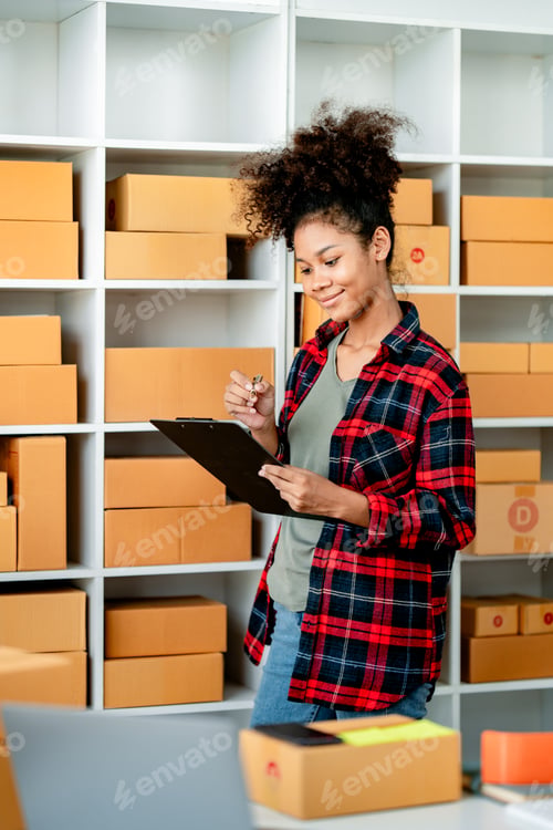 Preview: Young African woman prepares parcel boxes and checks online orders for delivery to customers on lapt