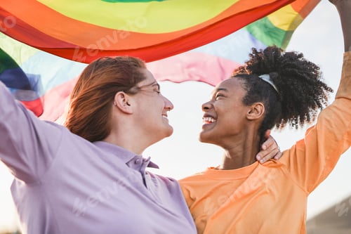 Vista previa: Happy gay female couple holding LGBT rainbow flag at pride parade