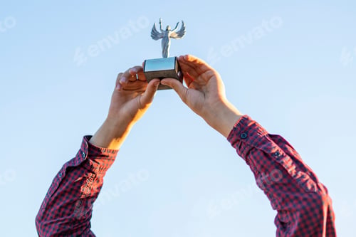 Preview: young person winning a competition, raised hands with a trophy figure against the sky with copy spac