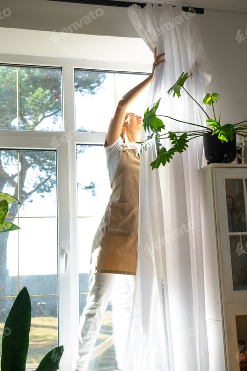 Preview: Woman in apron hangs transparent tulle curtains on large windows in the house inside