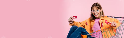 Preview: Panoramic concept of woman showing price tags with sale word while sitting in cart on pink