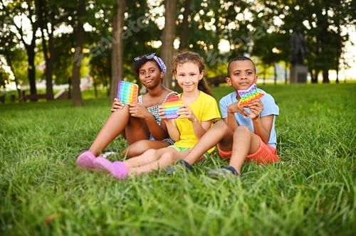 Preview: children of different races sit on the grass and play with an anti-stress toy pop-it