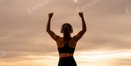 Preview: Rear view of athletic woman wearing headphones raising arms in victory gesture against sunset sky