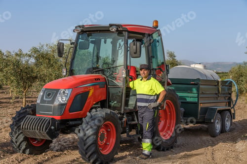 Preview: Farmer operating a red tractor with a trailer in an olive grove under a clear blue sky