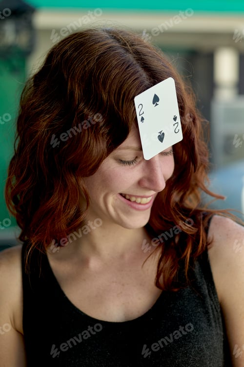Preview: A smiling woman with red hair has a playing card, the two of spades, stuck to her forehead.