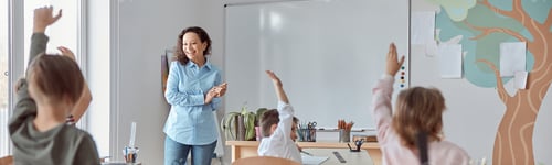 Preview: Young happy elementary school pupils at light modern classroom are rising their hands on a lesson