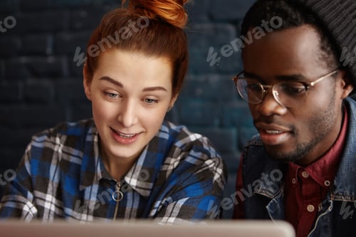 Preview: Cheerful Caucasian girl and African man in glasses and hipster hat watching video online, looking at
