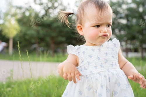 Preview: Toddler in a white dress plays in a green park, capturing childhood curiosity and innocence.