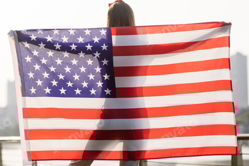 Preview: Happy young woman holding American flag outdoors on sunset. Portrait of woman from behind