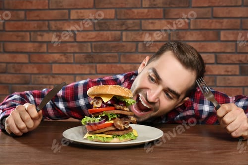Preview: Young hungry man with cutlery eating huge burger at table