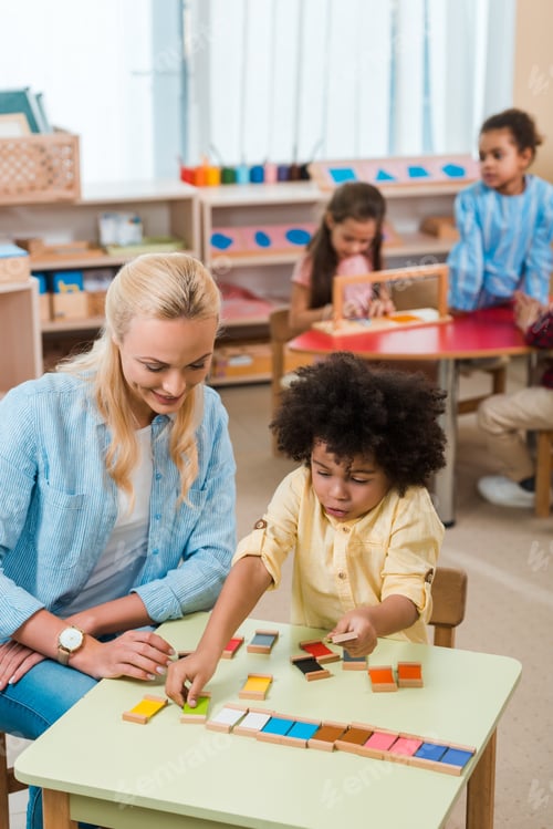 Preview: Selective focus of child playing game by smiling teacher with kids at background in montessori