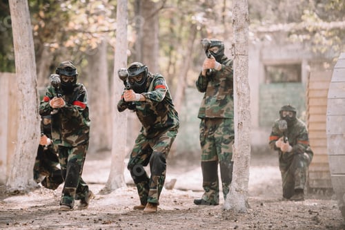 Preview: paintball team in uniform and protective masks running and aiming by paintball guns outdoors