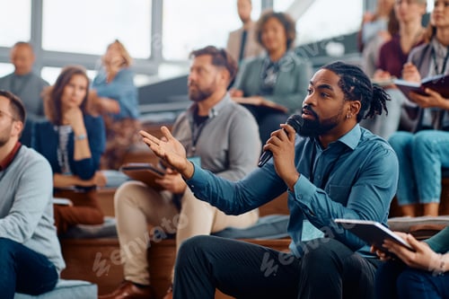 Preview: Black business seminar attendee asking a question while sitting in audience at convention center.