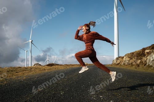 Preview: Young sportswoman jumping listening music near wind turbines