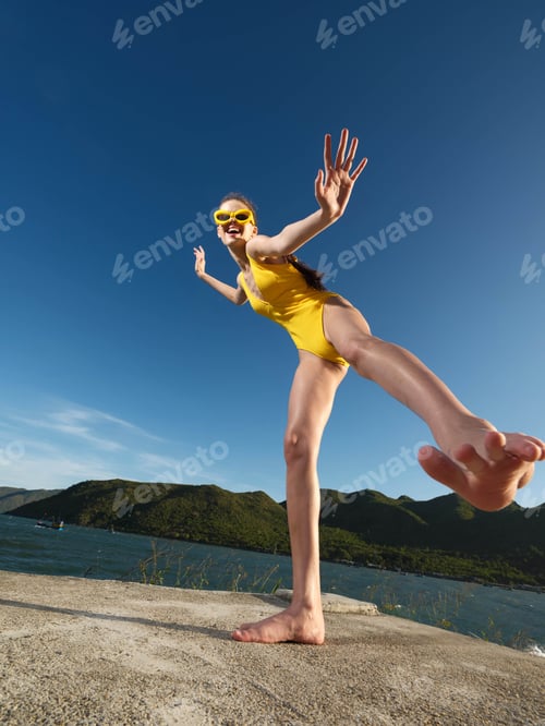 Preview: Yellow swimsuit woman standing on rocky edge near water and mountains in background, capturing a